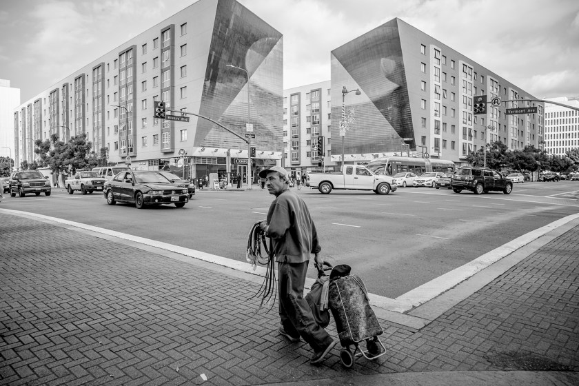 A man selling belts crosses the street at the corner of Vermont Avenue and Wilshire Boulevard across from the Vermont Metro Station. A luxury apartment complex surrounds the Metro Station entrance